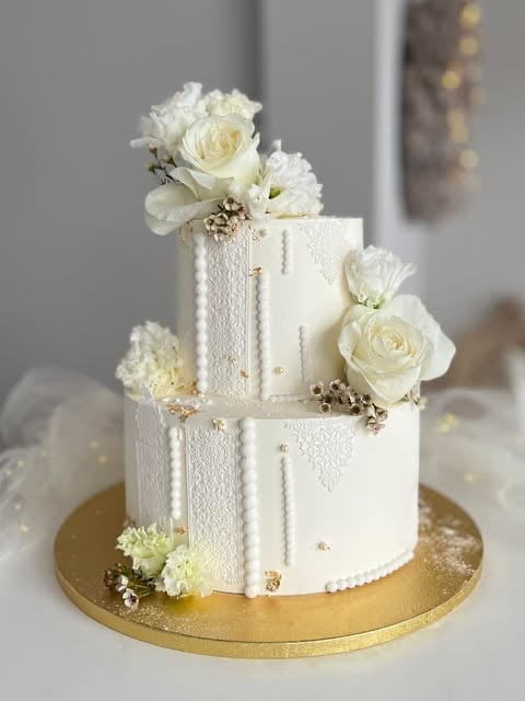 Three-tiered white wedding cake with textured frosting, gold base plate, and white roses with pearl accents