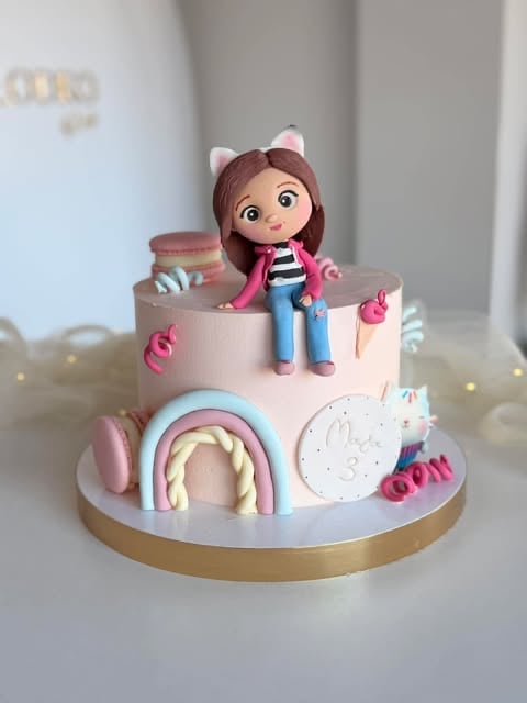 Two-tier pink and white decorated cake with a doll figurine on top, rainbow arch, decorative elements, and gold base on white background