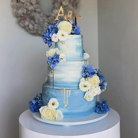 Four-tier blue and white wedding cake decorated with blue hydrangeas and white flowers, topped with a gold geometric cake topper