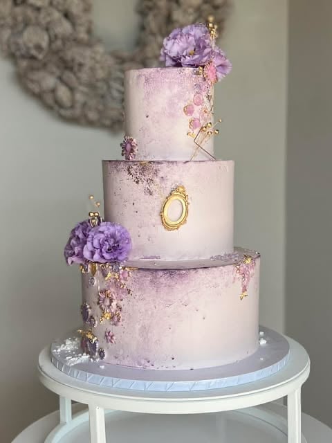 Three-tiered wedding cake with lavender frosting, gold accents, and purple peony flowers on a white cake stand