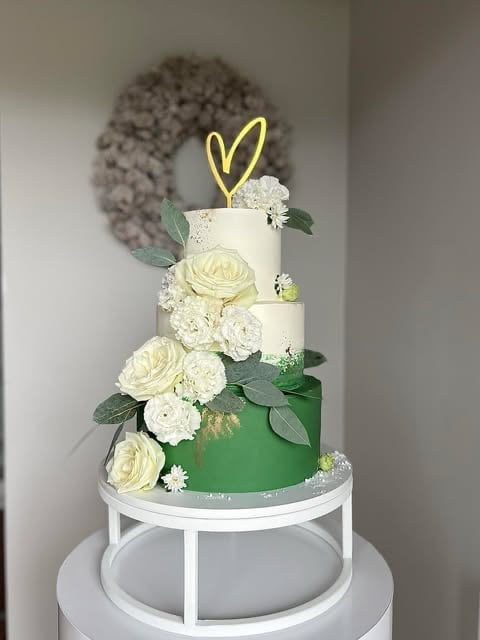 Multi-tiered wedding cake with white and green frosting, decorated with white flowers, eucalyptus leaves, and a yellow heart topper on a white cake stand