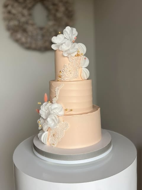 Three-tiered wedding cake with peach frosting, white flowers, and delicate lace details on a white base stand