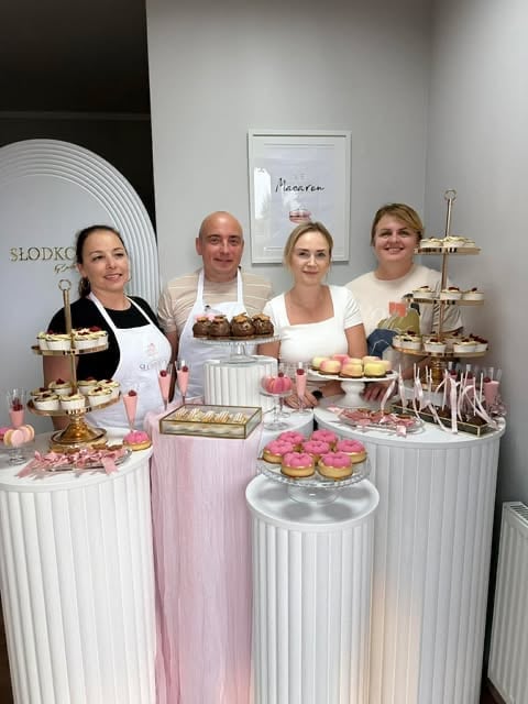 Four people standing behind a pink and white dessert display table with pastries, cupcakes, and tiered cake stands in an elegant bakery setting