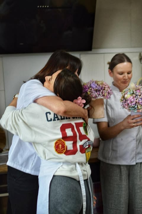Two women hugging while a third woman holds purple flowers, indoors near a display screen