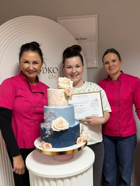 Three women in pink jackets posing with a three-tier blue and white wedding cake decorated with peach flowers, holding a certificate
