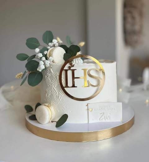 Elegant white tiered wedding cake with gold band, IHS monogram, fresh eucalyptus, white flowers, and cream-colored macarons