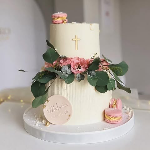 Elegant white tiered cake with gold cross, pink flowers, green foliage, and decorative macarons on a white cake stand