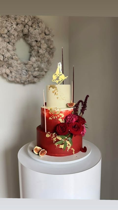 Three-tiered wedding cake with red bottom, cream middle, and gold leaf details, decorated with fresh red roses, figs, and tall dark candles on a white pedestal stand